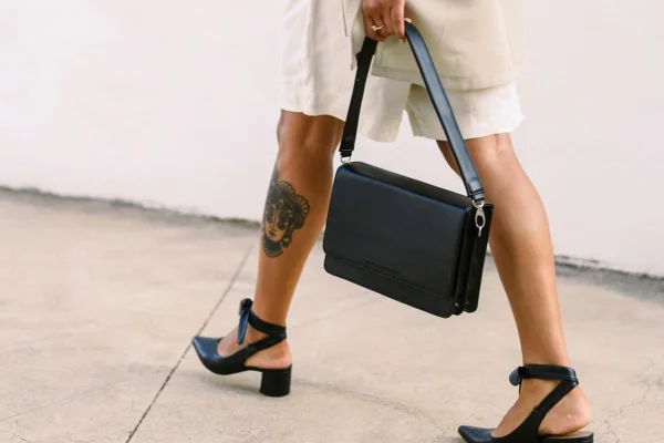 Mulher branca andando de salto alto preto, bolsa preta e short branco. Ela possui uma tatuagem na perna. Não é possível ver seu rosto, apenas a perna caminhando.