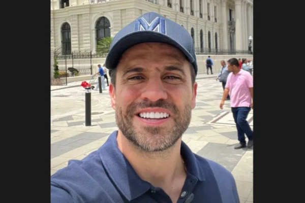 Imagem colorida de homem branco com barbar rala, com boné e camisa polo azul marinho. Ele faz selfie em frente a prédio antigo - Metrópoles