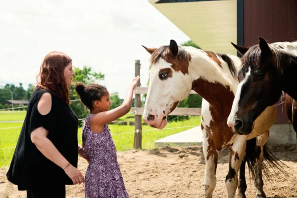 Imagem colorida de criança acariciando cavalo ao lado de uma pessoa adulta