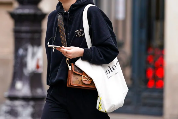 Pessoa andando com moletom preto, com broche da Chanel, e uma bolsa da Louis Vuitton. Ela também está usando uma ecobag.