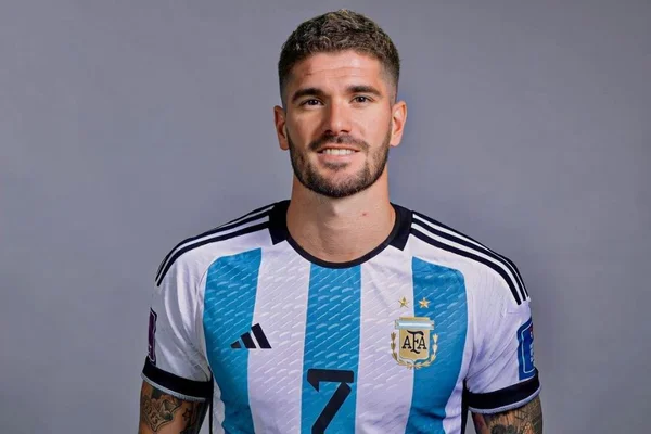 Foto do jogador Rodrigo de Paul em um fundo cinza e usando uma camisa da seleção argentina. Ele está sorrindo para a foto que mostra apenas do seu tronco para cima