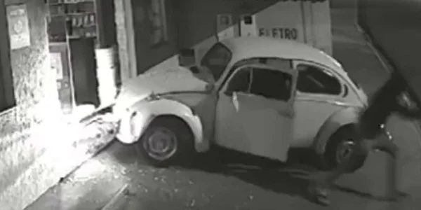 Imagem em preto e branco mostra homem vestido de preto saindo de um fusca que está estacionado na frente de uma loja com os vidros quebrados - Metrópoles
