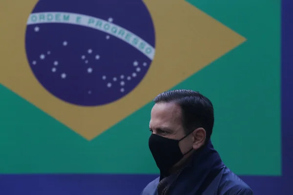 Governador de São Paulo, João Dória discursa de máscara em evento com bandeira do Brasil ao fundo. Ele segura um microfone e usa casasco - Metrópoles