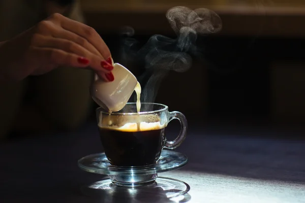 Foto colorida de uma mão feminina derrubando café quente em uma xícará que está enfatizada pela luz do ambiente - Metrópoles.
