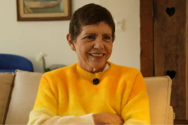 Imagem em cores mostra mulher branca, de cabelo curto com uma blusa amarela sorrindo e de braços cruzados