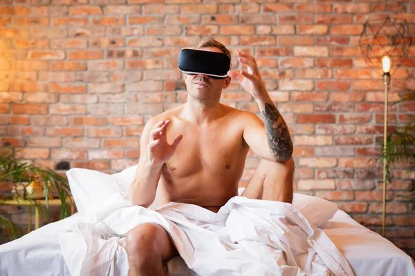 Foto colorida de homem nu coberto com lençol sentado na cama com óculos de realidade virtual - Metrópoles