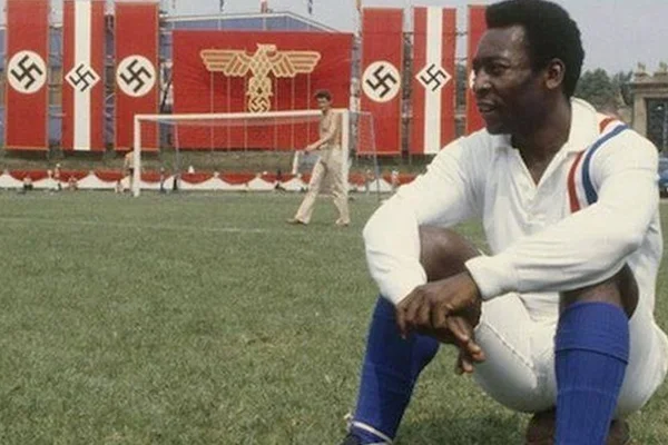 Cena do filme "Fuga para a Vitória" em que o craque Pelé aparece sentado em gramado de estádio com bandeiras nazistas ao fundo - Metrópoles