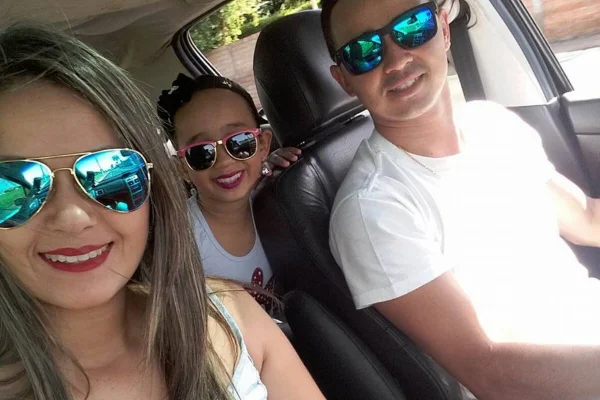 Em foto colorida mulher e homem, nos bancos dianteiros de carro, e menina no banco traseiro sorriem em selfie - Metrópoles