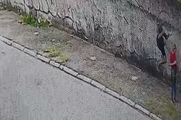 Imagens coloridas mostram homem de bermuda e boné escuros atingindo mulher de blusa vermelha que fala ao celular na numa calçada