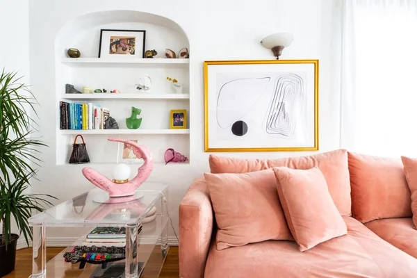 Sala decorada de forma moderna. É possível ver um sofá rosa, uma estante com livros e peças decorativas e um quadro na parede com moldura de madeira