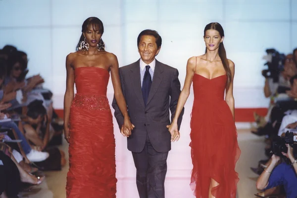 O estilista Valentino Garavani andando na passarela de um desfile ao lado das modelos Naomi Campbell e Gisele Bündchen. As duas usam vestidos vermelhos e ele usa terno e gravata. A foto é de 1999. - Metrópoles