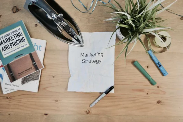 Na imagem há três livros, uma planta, um ferro de passar, alguns pinceis e um papel escrito Marketing Strategy - Metrópoles