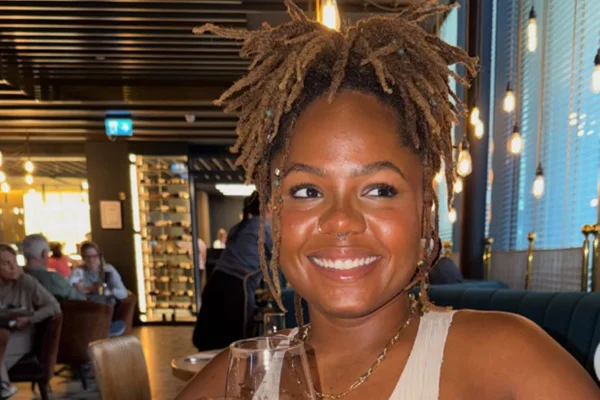 Foto colorida de mulher com cabelos amarrados, sorrindo para a foto em um ambiente de luz baixa - Metrópoles.