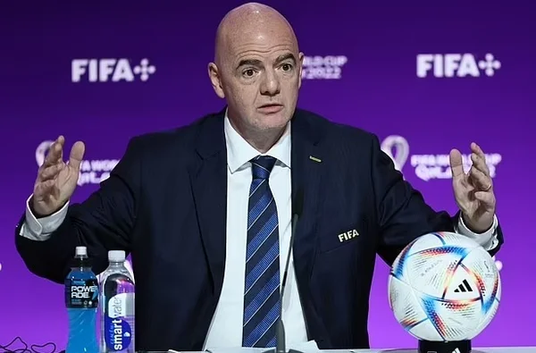 Foto de um homem careca e de terno na frente de um telão da Fifa falando com os braços abertos sobre a Copa do Mundo