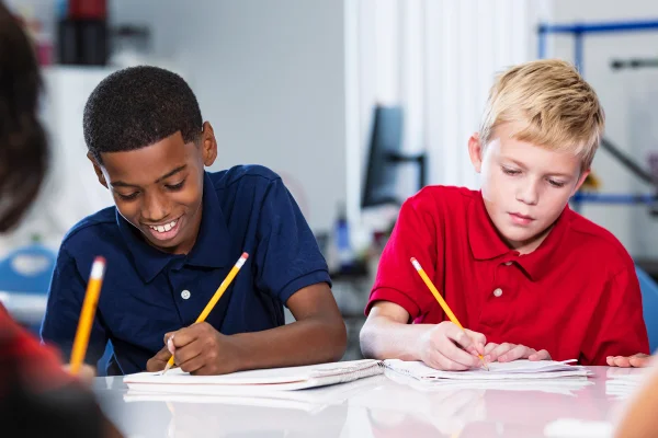 Alunos multirraciais do ensino médio escrevendo em sala de aula. Os dois alunos do ensino médio estão fazendo uma tarefa ou fazendo um teste. O menino afro-americano está sorrindo e seu colega tem uma expressão séria. Metrópoles