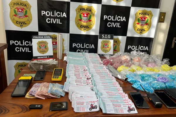Imagens coloridas mostram dinheiro, celulares, drogas e bilhetes de rifa clandestina apreendidos e expostos em mesa com banner da Polícia Civil de São Paulo ao fundo