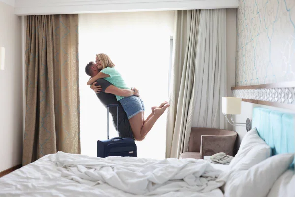 foto colorida de um casal se abraçando em um quarto de hotel com uma mala ao lado - Metrópoles