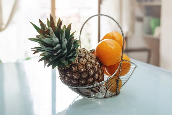 Cesta de metal com frutas - abacaxi e laranjas na mesa de vidro na cozinha. Metrópoles