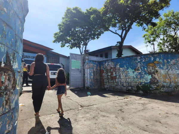 imagem colorida mostra mulher de mãos dadas com menina, estudante de uma escola. As duas estão de costas, andando em direção a uma escola - metrópoles
