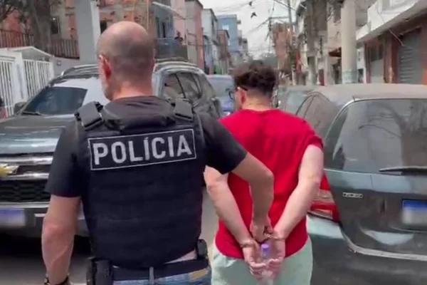 Imagem colorida de homem de camisa vermelha algemado com um policial atrás dele. Metrópoles