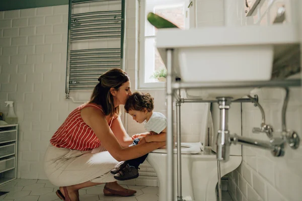 Foto mostra mãe ao lado de criança que se equilíbria em um vaso sanitário em um banheiro com tons claros - Metrópoles - mãe penico rotavirus diarreia criança