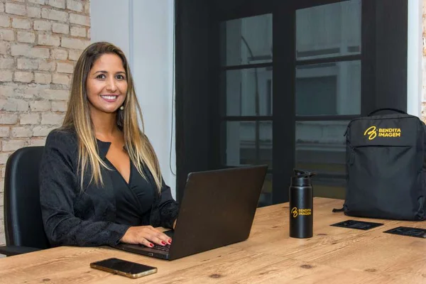 Na foto está Michele Barcena sentada em uma mesa que contém um notebook, um celular e uma garrafa de água - Metrópoles