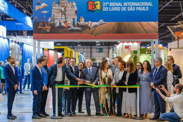Imagem colorida mostra homem branco grisalho, vestindo terno. Ele está ao centro de diversos homens e mulheres e segura uma fita em cores verde e amarela - Metrópoles