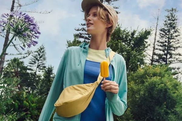 Em área verde, com árvores ao fundo, mulher usa roupa azul com boné e bolsa tiracolo amarela, da Uniqlo - Metrópoles
