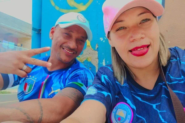 Casal vestido com camisetas azuis idêntica se cada qual usando boné. Esquerda, homem pardo, de bigodinho, sorri fazendo "v" de vitória com a mão esquerda. AO lado dele, mulher branca, loiras, olhos claros, mordendo a língua de forma marota com os dentes superiores - Metrópoles