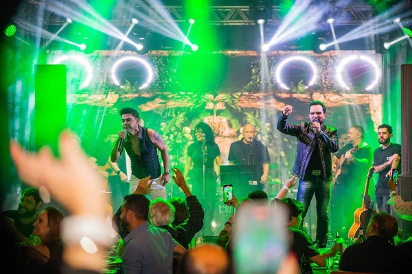 Zezé Di Camargo e Luciano cantando em palco com luzes verdes e público na frente - metrópoles