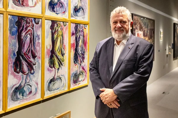 Carlos Vieira de braços cruzados ao lado de uma exposição de telas