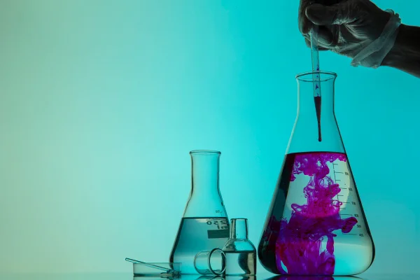 Foto colorida de tubos de ensaio, com fundo ver, líquido roxo e uma mão pingando algo no frasco, provocando uma reação química - Metrópoles