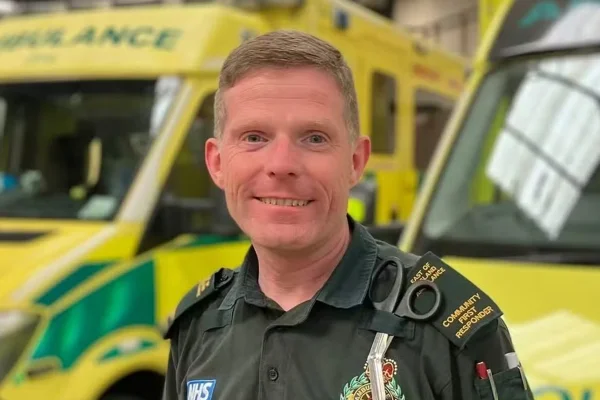 Imagem mostra Rhys Hibbert, que é um homem branco e loiro, sorrindo em frente a ambulâncias - Metrópoles
