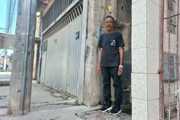 imagem colorida mostra homem em frente a sua casa; ele é negro, veste uma blusa cinza escura e uma calça jeans. ao seu lado está o portão de outra residência.