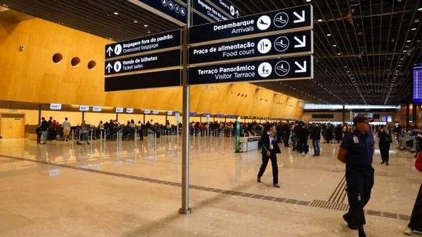 Placas em aeroporto com pessageiros passando ao fundo - Metrópoles