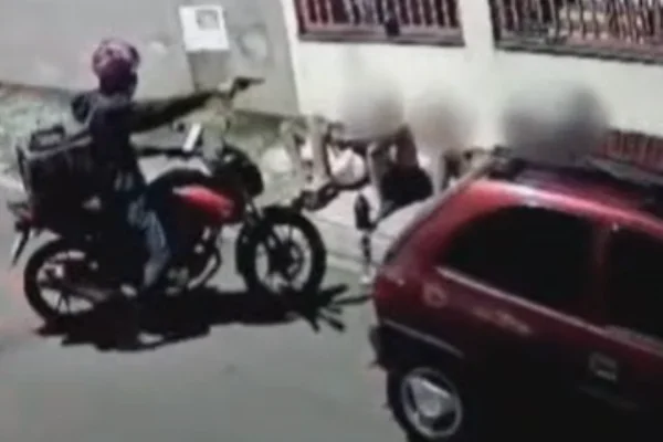 Imagem colorida de uma câmera de segurança mostra um motociclista armado apontando a arma para quatro jovens sentados em uma calçada - Metrópoles