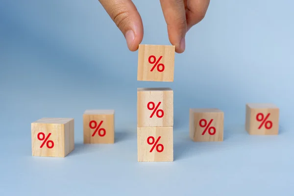 Imagem colorida de uma mão empilhando dados que exibem um símbolo de porcentagem na cor vermelha. Três dados estão empilhados uns sobre os outros e mais três estão separados, no chão - Metrópoles