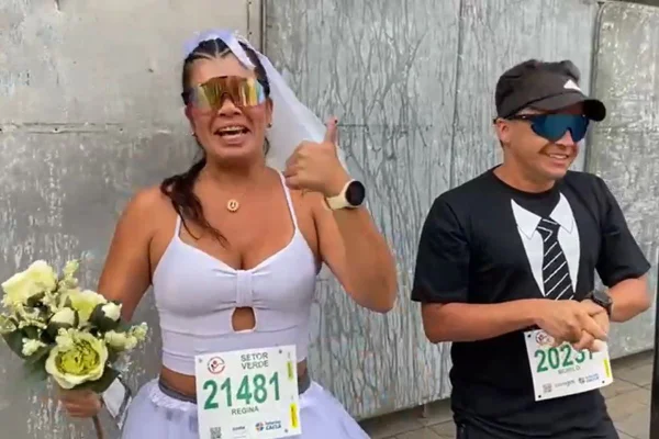 Imagem colorida de casal vestido de noivos. A mulher está à esquerda e segura um buquê. O homem, à direito, usa uma camiseta - Metrópoles