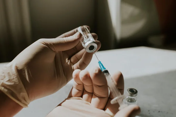 Uma mão usando luvas de látex segura um frasco com um líquido transparente e uma seringa. O profissional médico usa a seringa para extrair o medicamento do frasco de vidro. Metrópoles