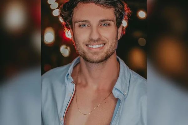 Na imagem colorida, um homem está posicionado no centro. Ele usa camiseta clara, calça jeans, tem cabelos curtos e loiros, e sorri para a câmera