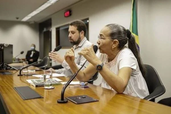 Fotografia colorida de mulher falando em microfone fixado em mesa com homem ao lado