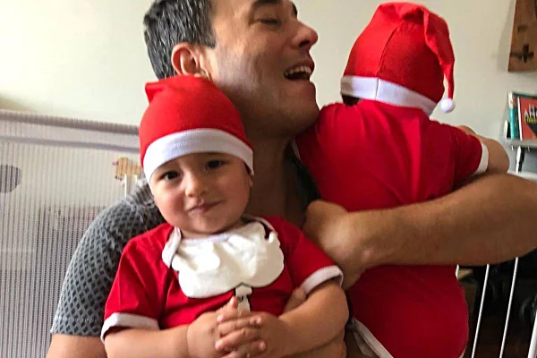 Na foto temos o jornalista André Rizek segurando os dois filhos bebes vestidos de body vermelho e gorro vermelho, um deles está de frente e o outro de costas