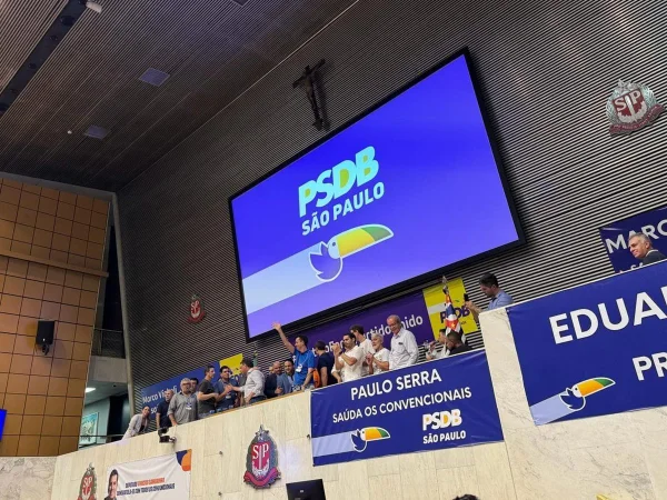 Imagem colorida mostra o plenário da Alesp com pessoas sorridentes e em pé na mesa diretora e uma série de faixas do PSDB