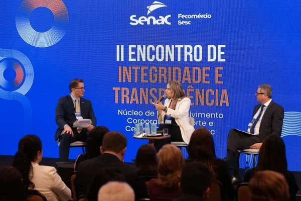 Auditório com plateia atenta a um painelista que fala no palco. Fundo azul com título do evento em destaque