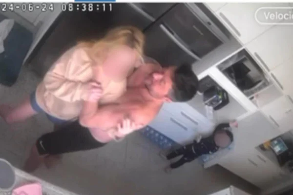 Homem branco sem camisa e de bermuda, preta, esgana mulher em cozinha, com criança olhando tudo ao fundo - Metrópoles