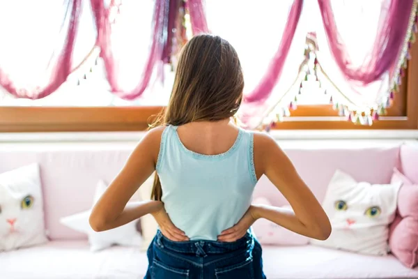 Imagem colorida mostra adolescente de costas, com as mãos na coluna, em quarto decorado em rosa - Metrópoles