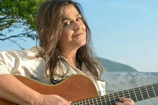 Foto da compositora Bia Bedran com um violão na mão. Ela usa blusa branca e está sorrindo timidamente - Metrópoles
