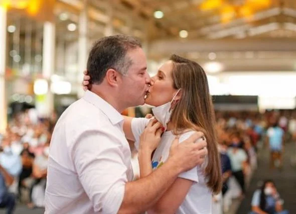 O governador de Alagoas e pré-candidato ao Senado, Renan Filho, beija frente a uma multidão sua esposa, Renata Calheiros. Eles estão de perfil e usam roupas brancas - Metrópoles