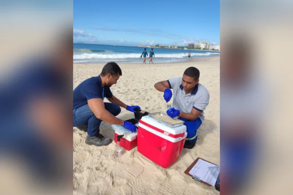 Dois funcionários da prefeitura do Rio de Janeiro fazem uma análise com ferramentas específicas da areia de uma praia. Ao fundo, o mar e pessoas caminhando. Praia de Botafogo está própria para banho após anos poluída - Metrópoles
