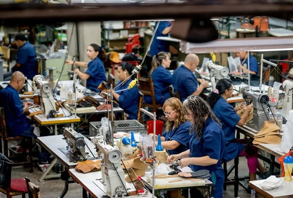 Mulheres e homens trabalhando em uma fábrica de roupas. Eles mexem em máquinas de costura e vestem, todos, um uniforme que é um macacão azul reoneração